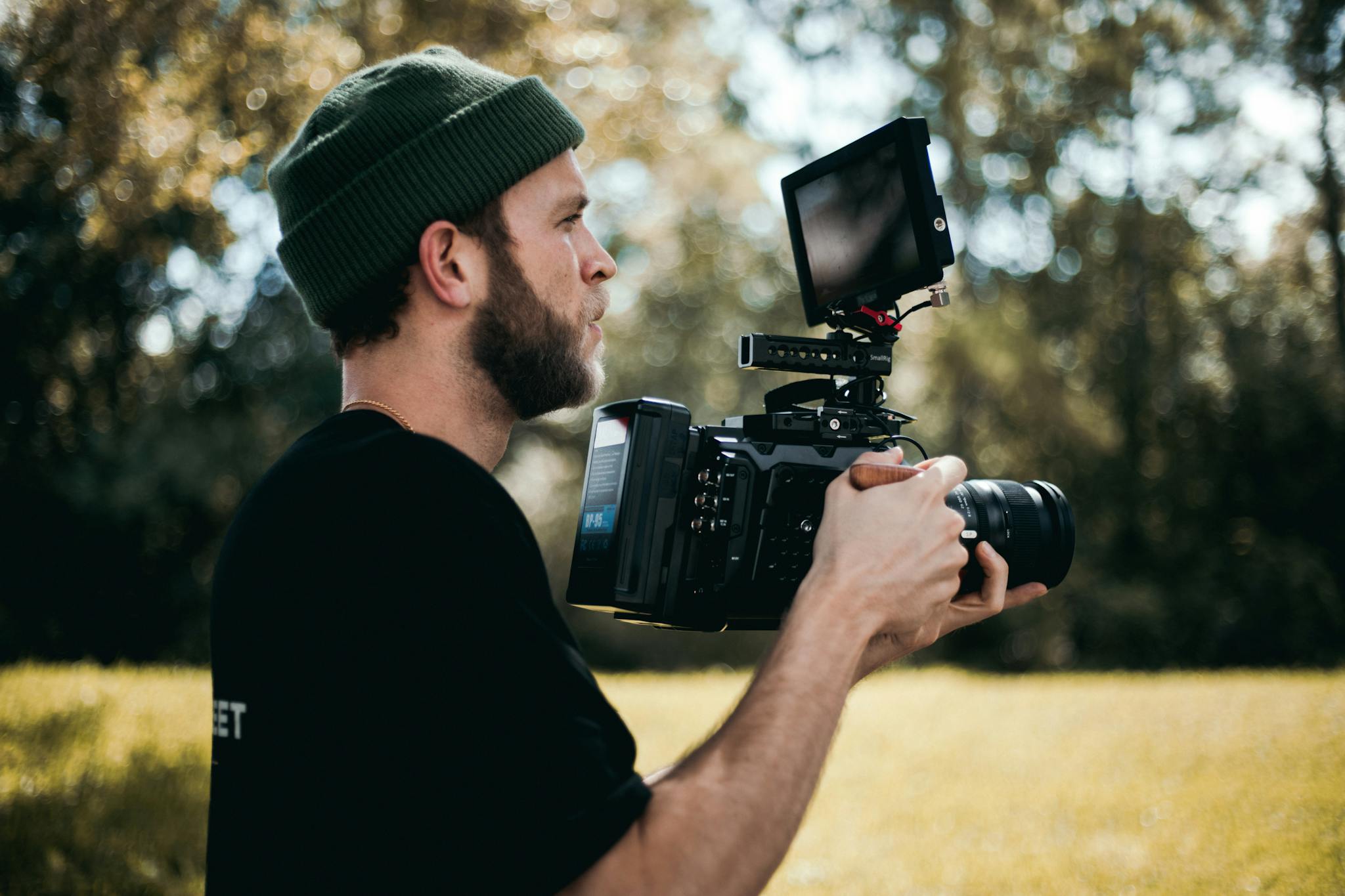 A bearded man wearing a beanie operates a professional camera outdoors, capturing a scene in nature.